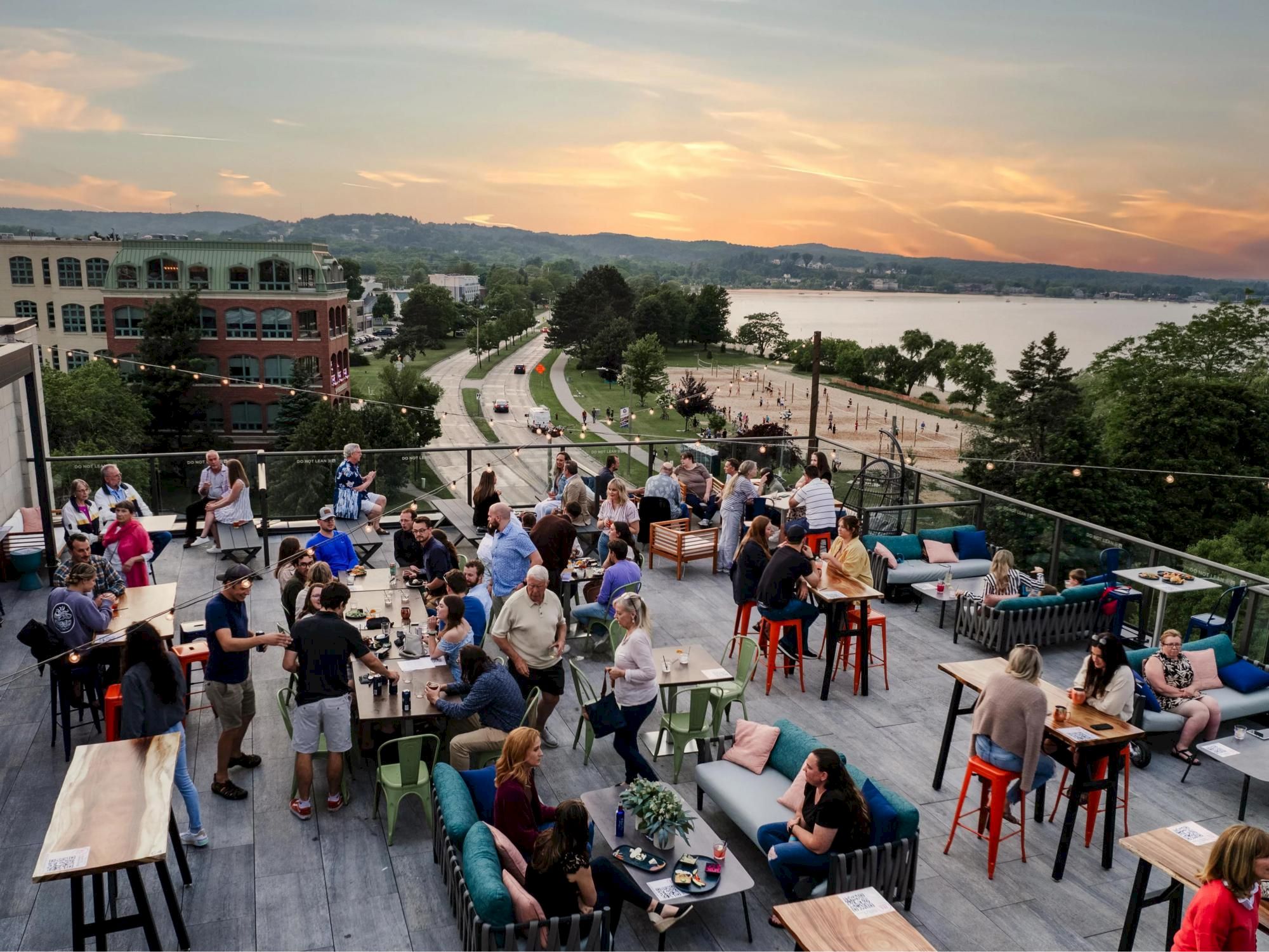People are enjoying a rooftop terrace with tables and seating at sunset, overlooking a road, park, and body of water in the background.