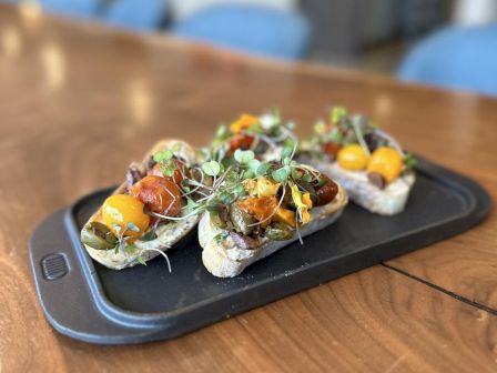 The image shows a platter with several pieces of bruschetta topped with roasted tomatoes, greens, and other toppings on a wooden table.