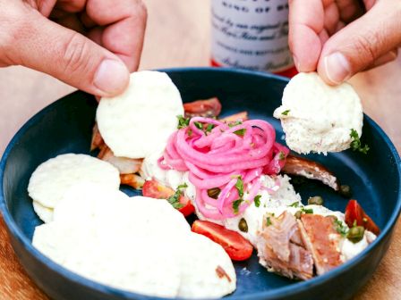 A plate with crackers, pickled onions, and meat, held by two hands. A beverage can is partly visible in the background.