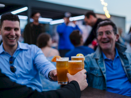 People are gathered outdoors at a table, raising glasses of beer for a toast under string lights in a lively atmosphere.
