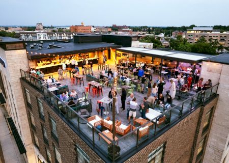 A vibrant rooftop bar scene with people socializing, tables, and seating, set atop a building overlooking a cityscape during the evening.