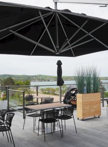 A rooftop patio with seating, tables, blue chairs, and large black umbrellas overlooks a scenic landscape with a road and greenery.
