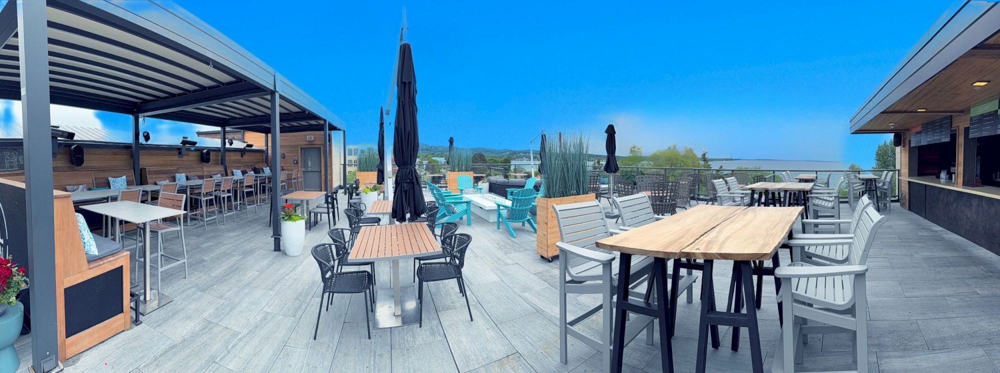 A spacious, modern outdoor rooftop seating area with tables, chairs, umbrellas, and a view of a calm landscape under a clear blue sky.