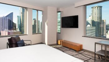 The image shows a modern hotel room with a large bed, a flat-screen TV, a cityscape view through multiple windows, and stylish furniture.