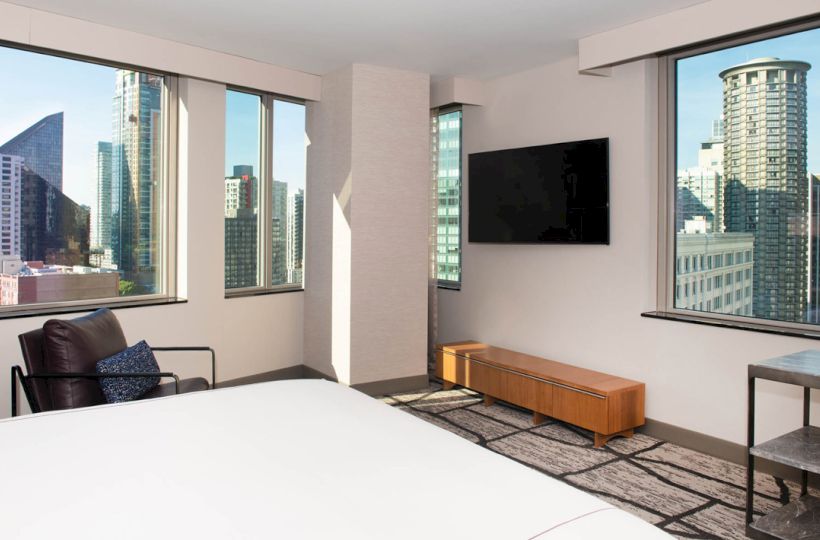 The image shows a modern hotel room with a large bed, a flat-screen TV, a cityscape view through multiple windows, and stylish furniture.