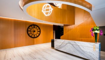 The image shows a modern lobby with a marble reception desk, a spiral wooden ceiling, a decorative clock, and hanging lights.