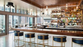 The image shows a modern bar with stylish barstools, a well-stocked liquor shelf, and large windows overlooking a city view.
