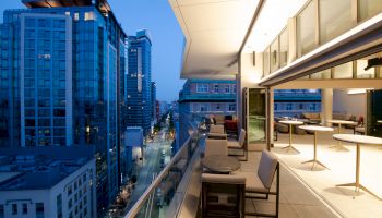 A modern rooftop terrace with seating overlooks a cityscape of tall buildings during dusk, under a clear blue sky.