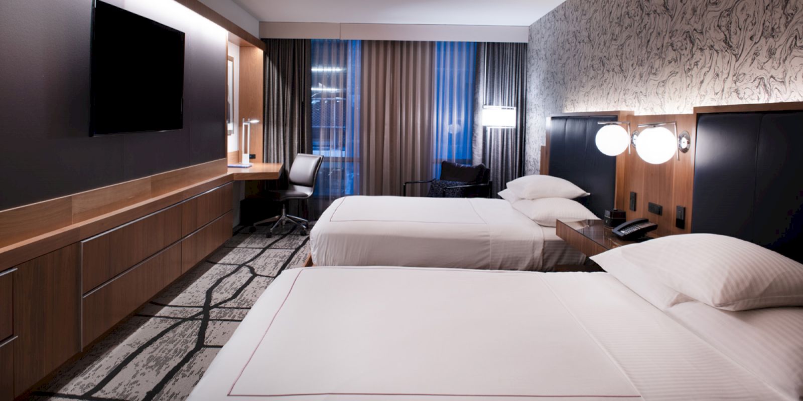 The image shows a modern hotel room with two beds, a flat-screen TV, a desk, and a patterned carpet and wall covering, softly lit.