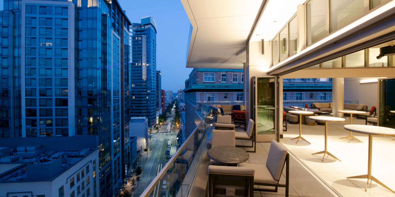 A modern rooftop terrace with seating overlooks a city street lined with tall buildings at dusk.