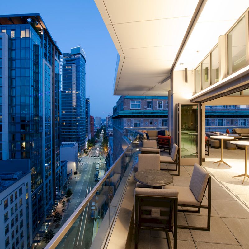 A cityscape view from a modern balcony at dusk, showing outdoor seating and high-rise buildings under a deep blue sky.