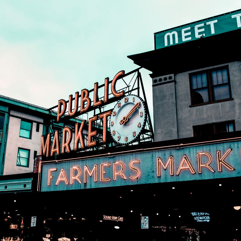 The image shows a neon sign for "Public Market" and "Farmers Market" with a clock, and nearby buildings under a cloudy sky.