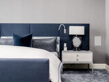A modern bedroom with a navy bed, white bedding, two-tone pillows, and a nightstand with a lamp and decor against a gray wall.