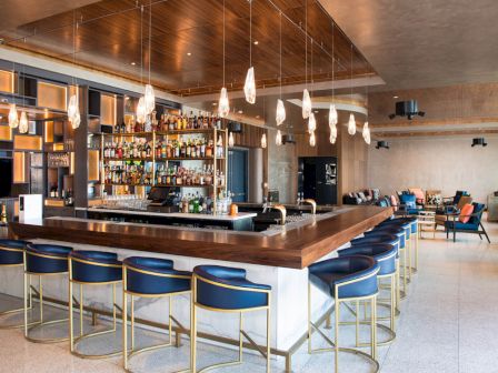 A modern bar with a central counter, blue stools, hanging lights, and shelves stocked with bottles, surrounded by seating and decor.