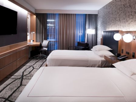 A modern hotel room with two beds, a flat-screen TV, desk, and lamp. Neutral tones and large window with curtains complete the look.