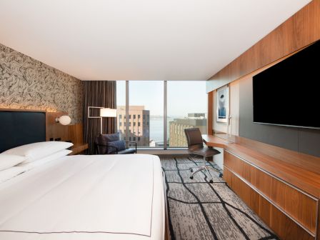 A modern hotel room with a large bed, desk, and TV, featuring a city view through wide windows ending the sentence.