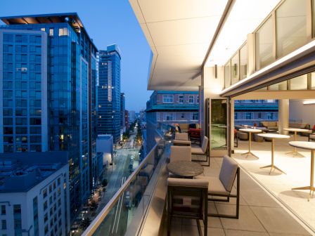 The image shows a modern rooftop terrace with seating and cityscape views, featuring tall buildings and a lit, contemporary interior.