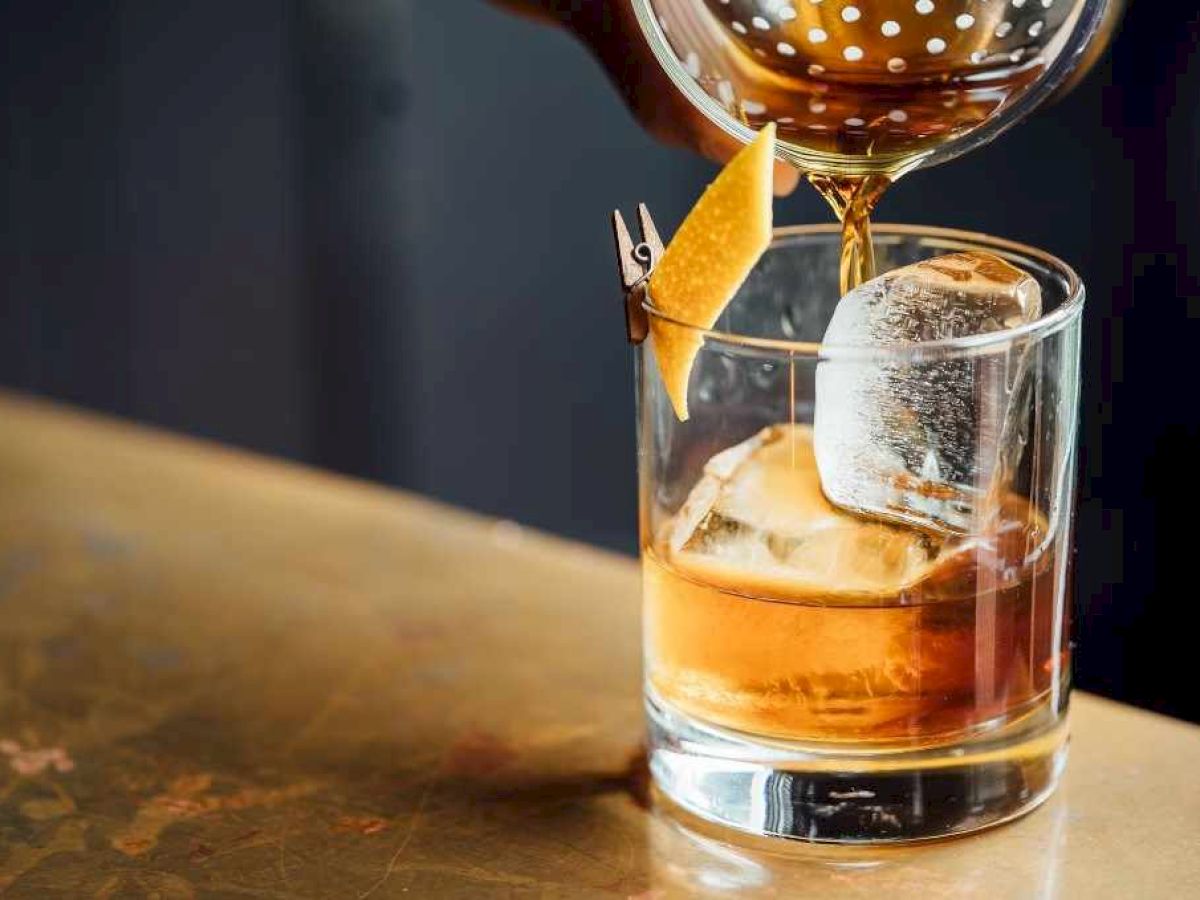 A drink is being poured into a glass with ice and a lemon twist garnish, sitting on a table.