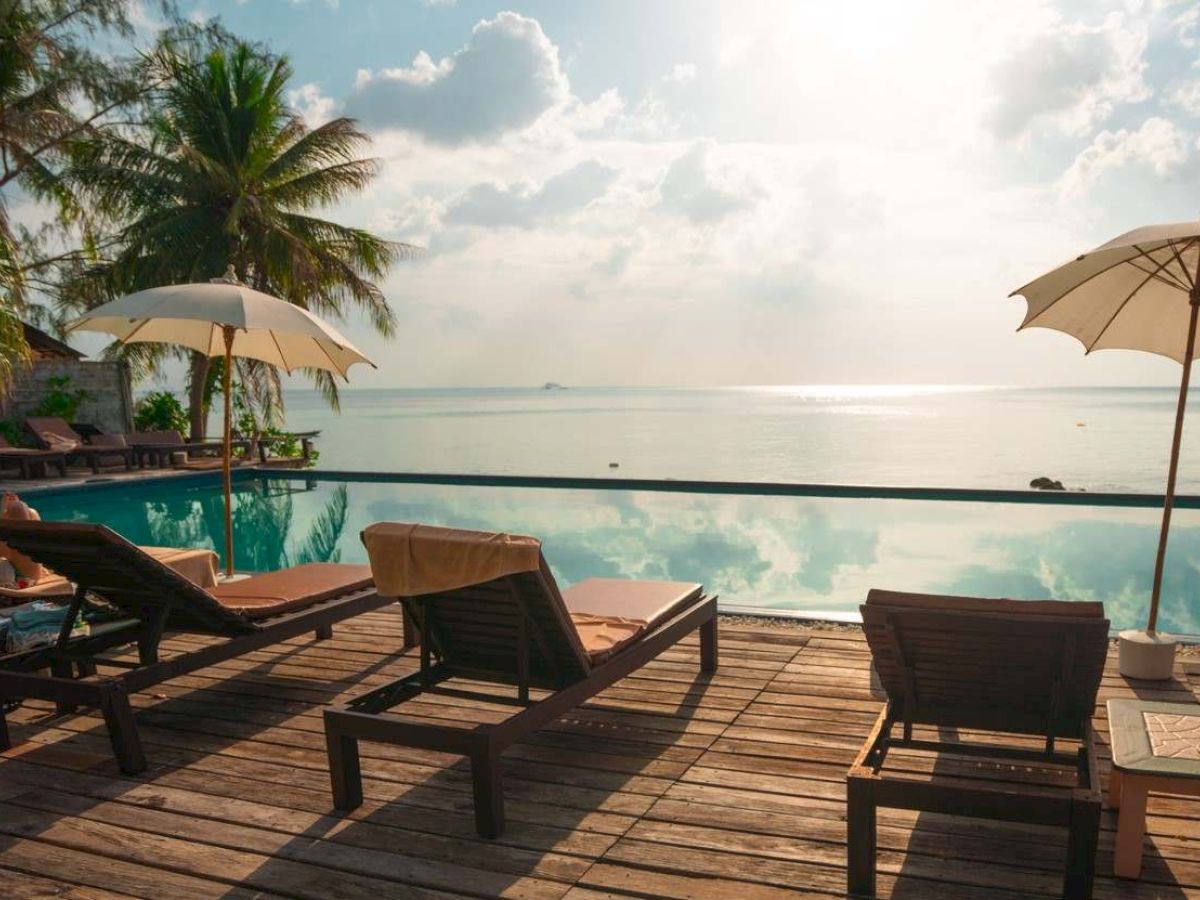 Beachside pool with lounge chairs, umbrellas, and scenic ocean view under a sunny sky, surrounded by lush palm trees.
