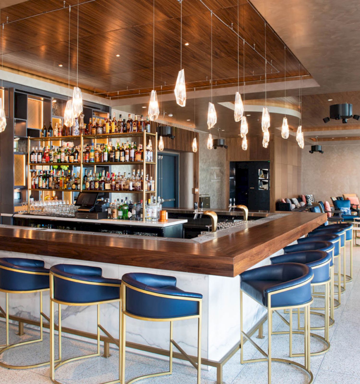 A modern bar with a stocked back wall, elegant hanging lights, and blue cushioned stools around a wooden counter in a stylish setting.