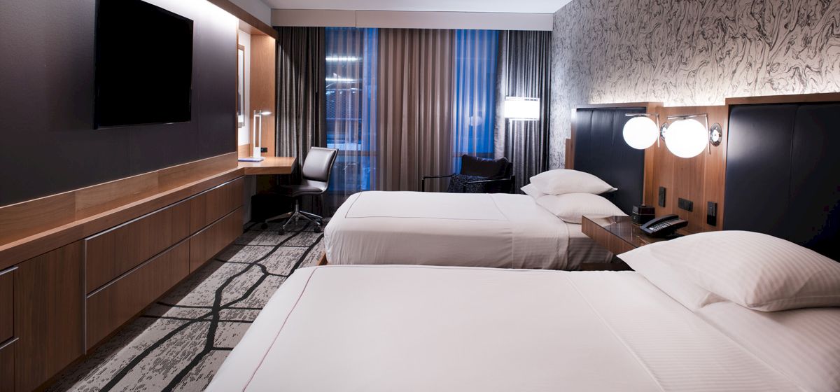 A modern hotel room with two beds, a flat-screen TV, a desk with a chair, and stylish decor, featuring neutral tones and ambient lighting.