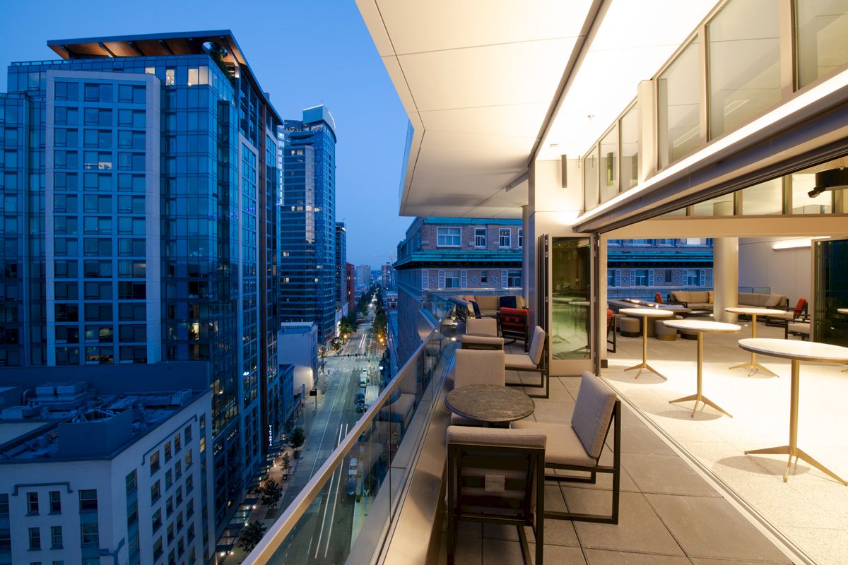 A cityscape at dusk features a modern balcony overlooking tall buildings and a street, with seating and warm interior lighting visible.