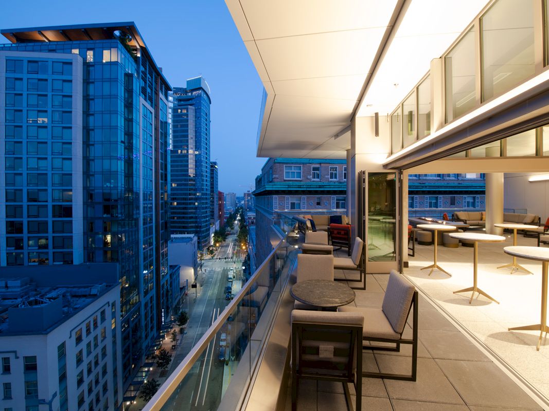 A cityscape at dusk features a modern balcony overlooking tall buildings and a street, with seating and warm interior lighting visible.