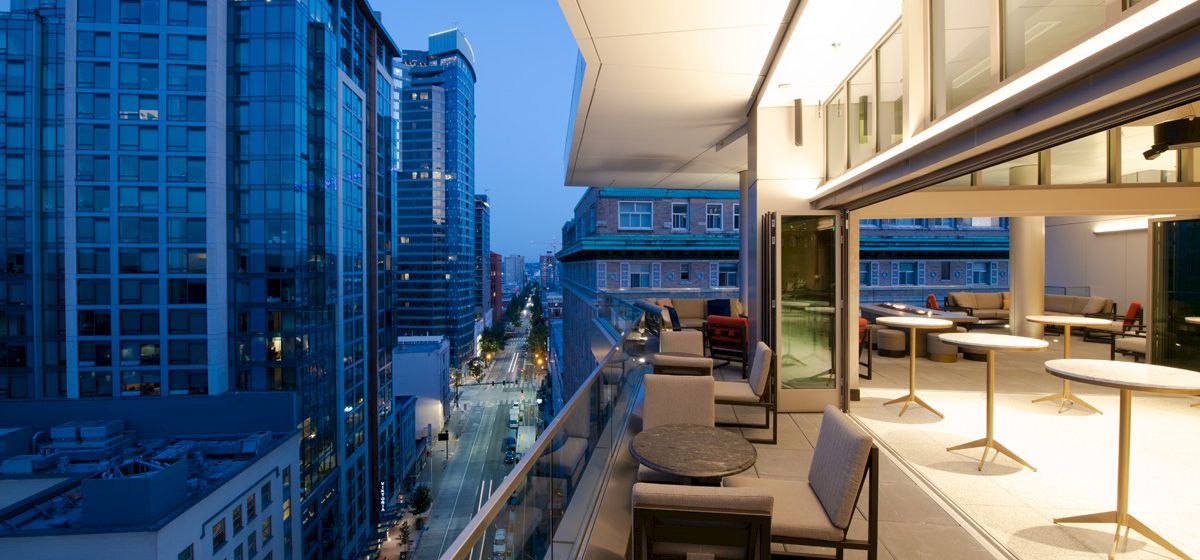 A cityscape at dusk features a modern balcony overlooking tall buildings and a street, with seating and warm interior lighting visible.