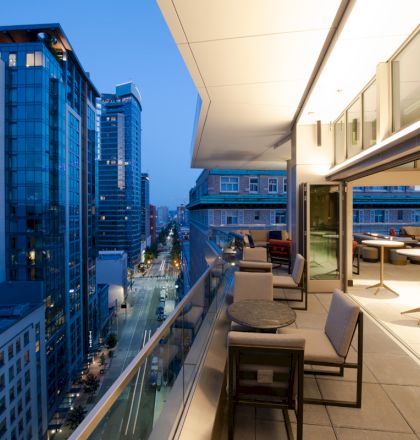 A modern cityscape with tall buildings and a stylish outdoor terrace at dusk, featuring seating and tables under warm lighting.