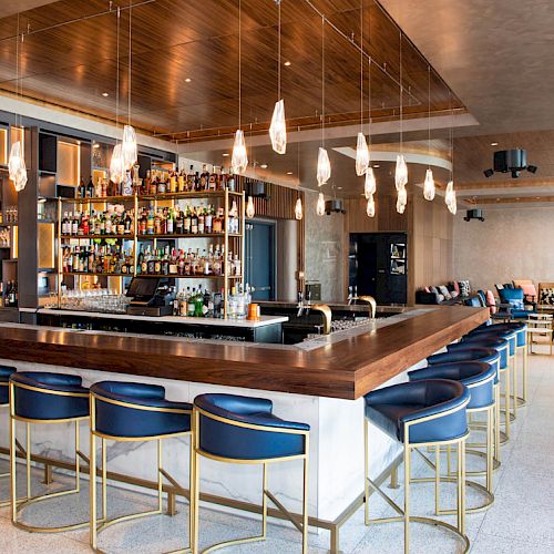 The image shows a modern bar with a wooden countertop, blue bar stools, hanging lights, and a stocked liquor shelf in a spacious lounge area.