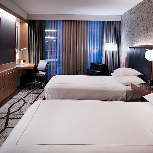 The image shows a hotel room with two beds, a wall-mounted TV, a desk with a chair, and stylish decor and lighting accents.