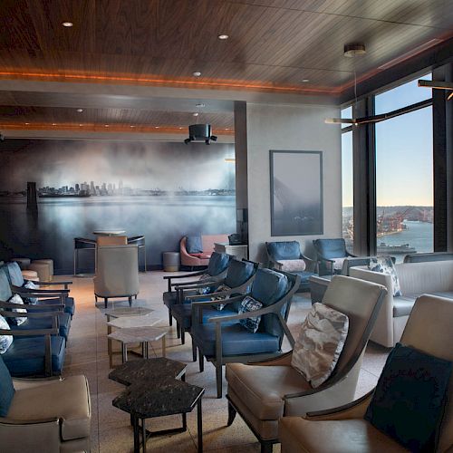 A modern lounge with numerous chairs and tables, a large window view of the sea and a cityscape mural on the wall.