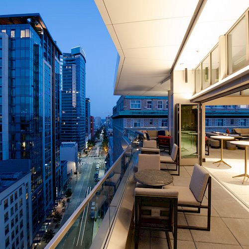 A modern rooftop terrace overlooks a city street lined with tall buildings, featuring outdoor seating and warm evening lighting.