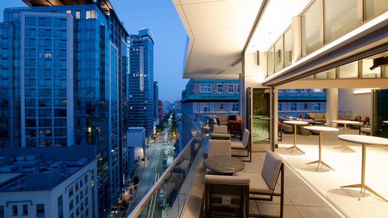 Rooftop terrace with seating, overlooking a cityscape with tall buildings and illuminated streets at dusk.
