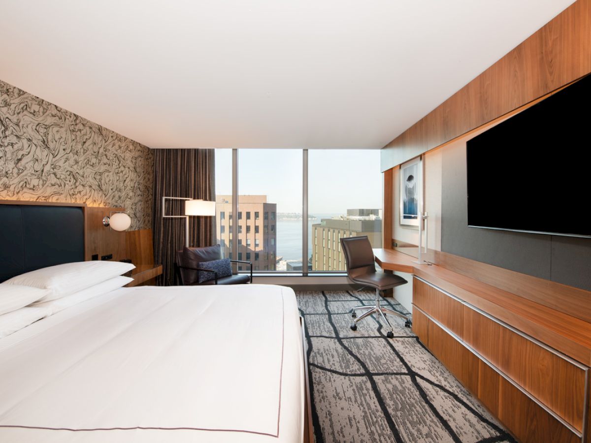 A modern hotel room features a large bed, wall art, a desk with a chair, a big flat-screen TV, and a view of city buildings and water.