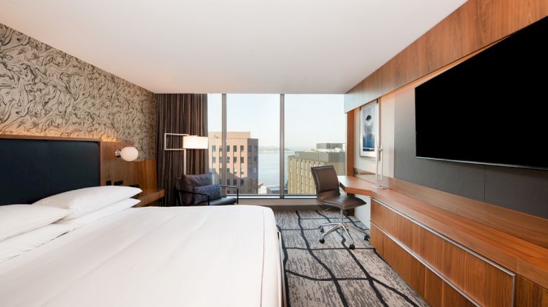 A modern hotel room features a large bed, wall art, a desk with a chair, a big flat-screen TV, and a view of city buildings and water.