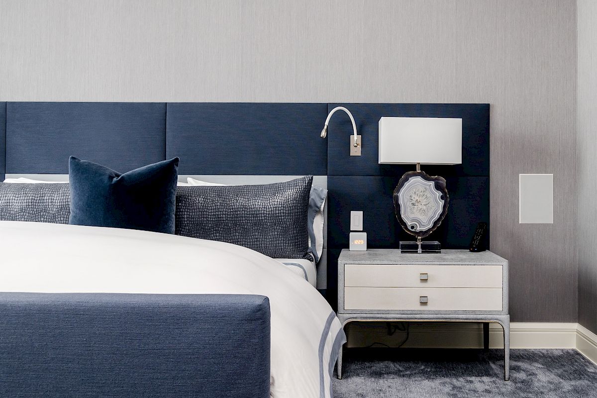 A modern bedroom with a blue upholstered bed, gray pillows, a white nightstand, a decorative lamp, and a reading light.