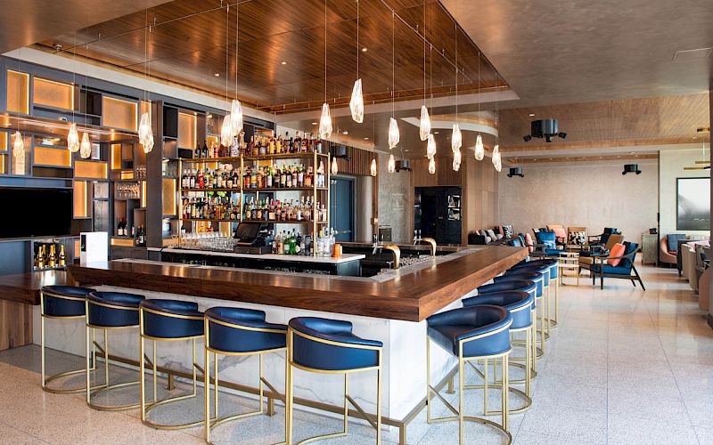 The image shows a modern bar with sleek lighting, a stocked back wall, and blue chairs around a wooden countertop.