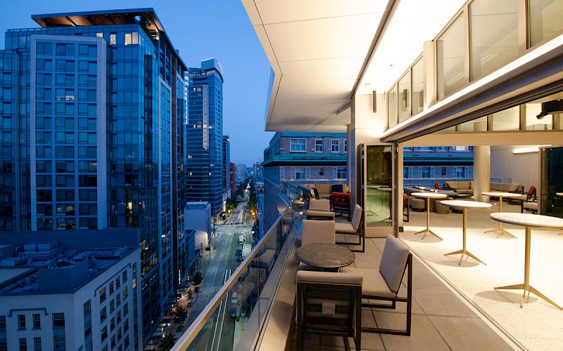 A modern balcony view at dusk with city skyscrapers, outdoor seating, and warm interior lighting.