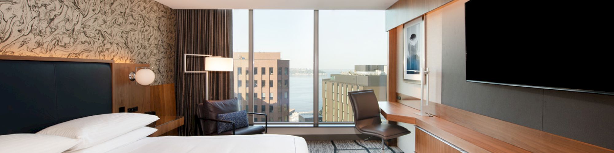 A modern hotel room features a bed, wall art, desk, chair, large TV, and a window with a city view, creating a cozy and stylish space.
