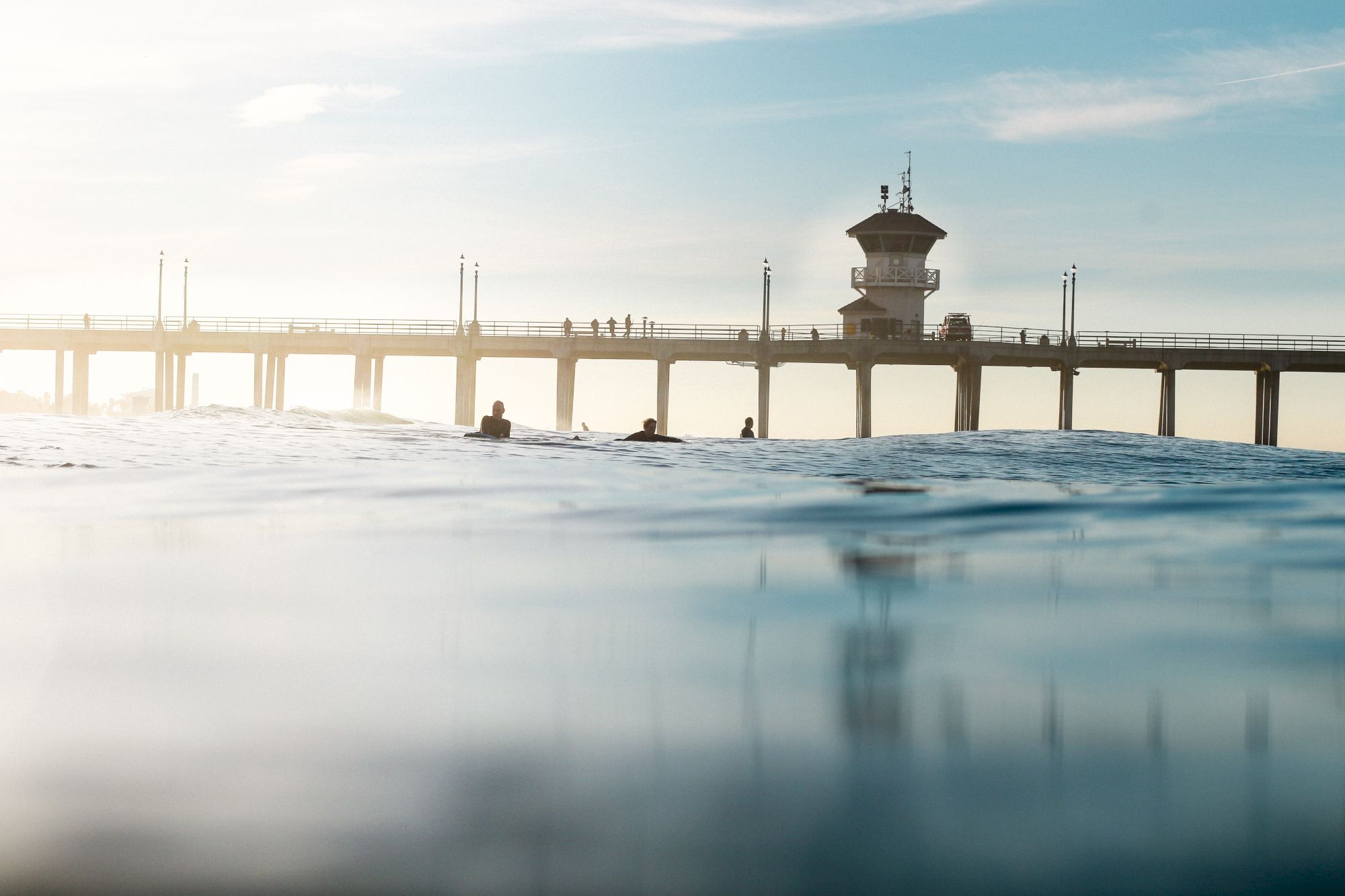 The image shows a pier extending over the ocean with several people on it, and surfers are visible in the water below.